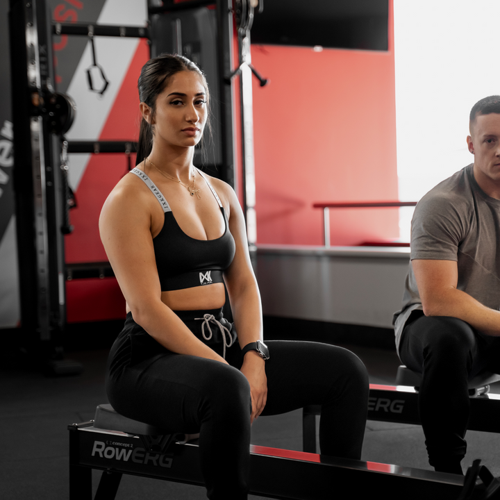 Female model wearing IXK Gear Cropped Trackies in Black paired with IXK X Sports Bra in Black. Gym background.